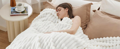 Een vrouw slaapt rustig onder een comfortabele Act Design Shop Comfortdeken 150x200cm op een bed met beige kussens. Naast haar staat een tafeltje met een kopje koffie en een boek met de titel ‘The.