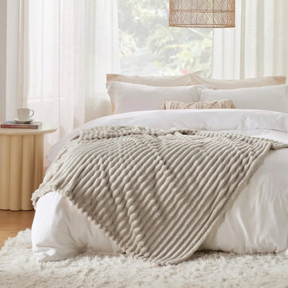 Een gezellige slaapkameromgeving met een zacht wit bed bedekt met een zachte, beige "Act Design Shop Comfort Deken 150x200cm" deken. Zonlicht filtert door transparante gordijnen naast een weelderig groen uitzicht door het raam.